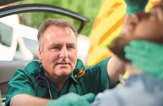 two ambulance paramedics stabilise a car crash victim. They are wearing green ambulance uniform typical of uk paramedics. One is sitting in the back of the car stabilising the victim's head whilst the other paramedic is assessing the casualty . The scene is shot through the car window.